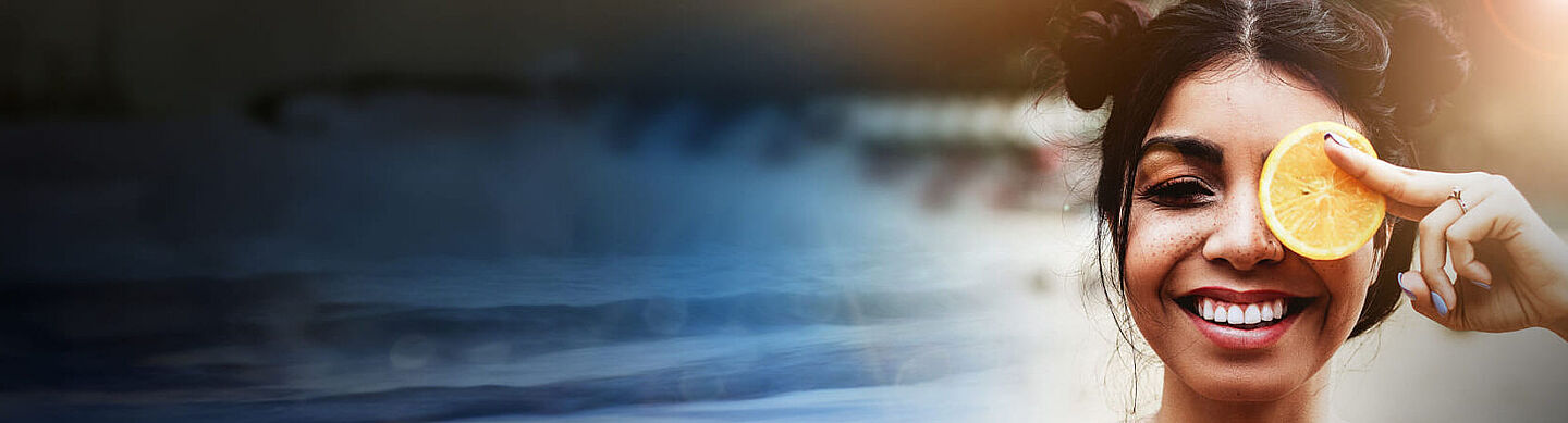A laughing woman holds a slice of lemon in front of her left eye