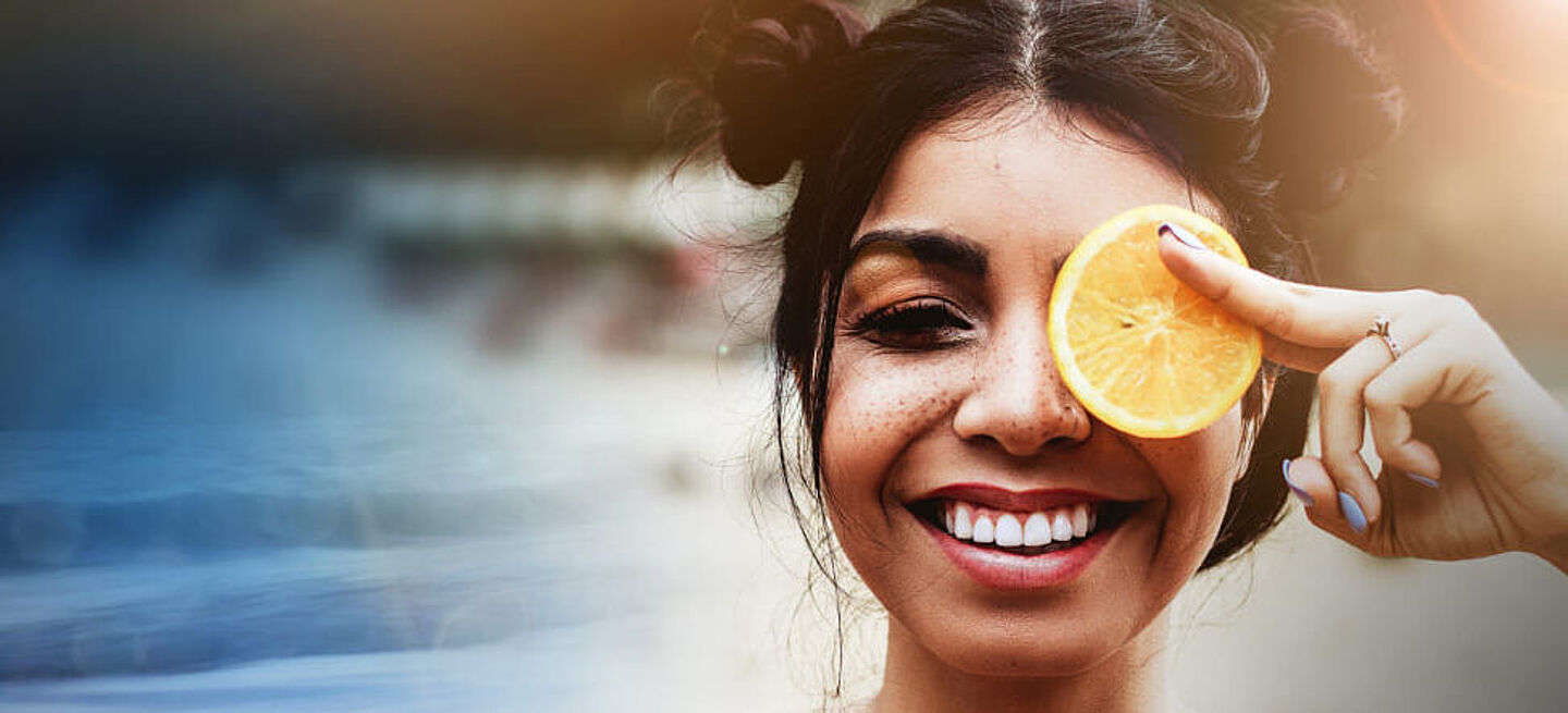 A laughing woman holds a slice of lemon in front of her left eye
