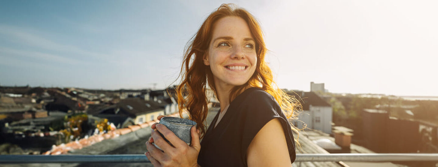 Lächelnde Frau, die einen Becher in den Händen hält und auf einer Dachterrasse steht. Die Szene ist von goldenem Sonnenlicht geprägt; dahinter erstreckt sich ein urbaner Stadtblick. Das Motiv vermittelt Genuss, Erfrischung und einen modernen Lifestyle.