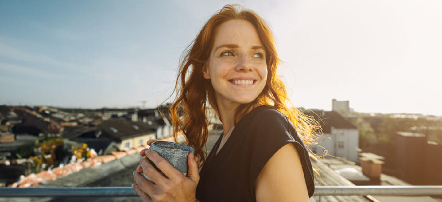 Lächelnde Frau, die einen Becher in den Händen hält und auf einer Dachterrasse steht. Die Szene ist von goldenem Sonnenlicht geprägt; dahinter erstreckt sich ein urbaner Stadtblick. Das Motiv vermittelt Genuss, Erfrischung und einen modernen Lifestyle.