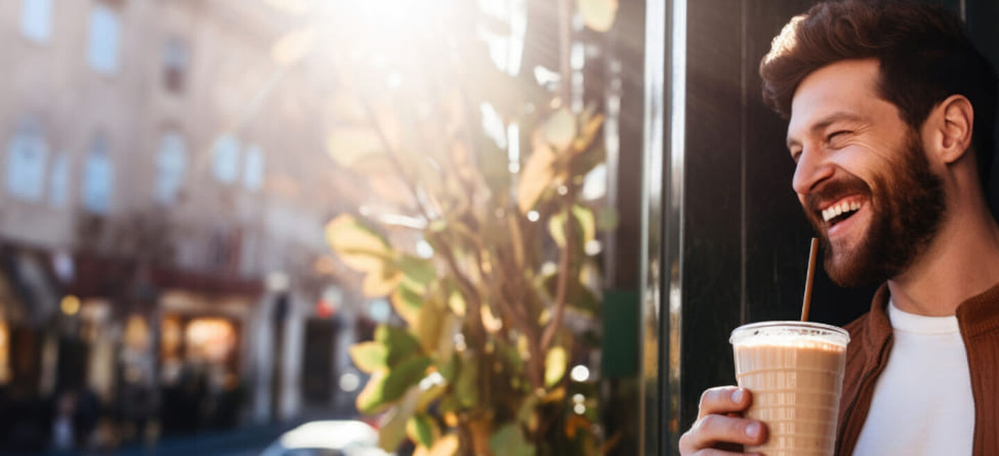 Bir adam şehirde bir kahve-to-go (buzlu kahve/soğuk kahve) içiyor. Sıcak ışık ve bulanık kafe arka planı, modern kahve keyfi ve yaşam tarzını yansıtıyor.